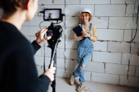 Photographer shooting a happy female construction engineer in a white helmet. Over a rough gray brick wall.の写真素材