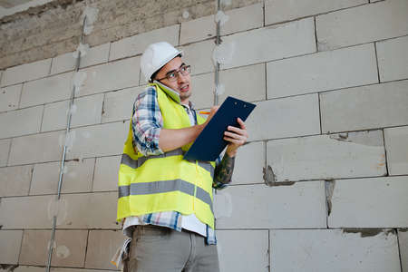 Overworked construction engineer in a white helmet discussing problems on the phone, writing it down on a clipboard. Standing next to a rough gray brick wall.の写真素材