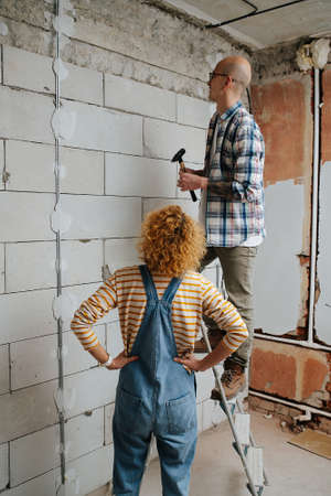 Busy couple making new apartment overhaul together, looking at the wall. Man is standing on a stepladder, woman has hands on her sides.の写真素材