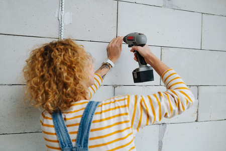 Behing view of a female construction worker drilling gray brick wall with electric screwdriver.の写真素材