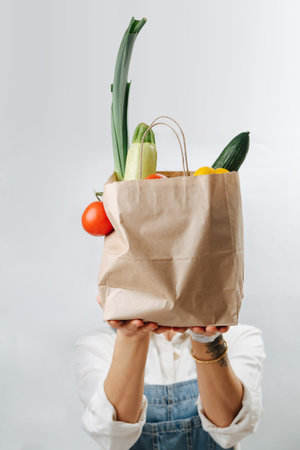 Blocking her face woman holding a paper bag filled with colorful vegetables
