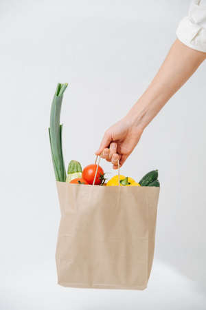Hand holding grocery paper bag filled with colorful vegetables over white background. Eco friendly compostable products.の写真素材