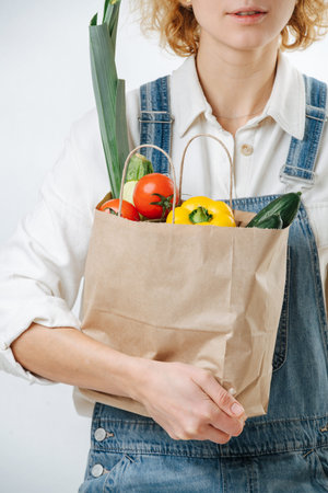 Cropped image of a woman holding a paper bag filled with colorful vegetables over white background....