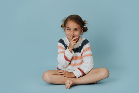 Gossipy little girl in striped sweater sitting cross-legged on the floor over blue background. She is pressing index finger to her lips. looking at the camera.の写真素材