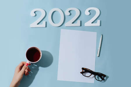 Top view of a blank sheet, 2022 number, glasses, pen and a hand holding a mug. Flat lay, new year composition. On a blue surface.の写真素材