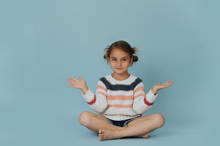 Sitting in meditation easy pose little girl in striped sweater on the floor over blue background. Looking with a smile at someone.の写真素材