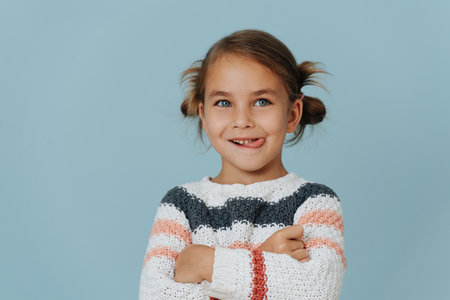 Perky little girl in striped sweater standing with hands crossed, sticking out tongue over blue background.の写真素材