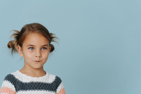 Resentful tight-lipped little girl in striped sweater, hair in buns over blue background. looking at...