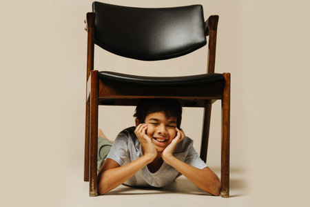 Carefree indian boy playing with an armchair lying under it with eyes closed, leaning on his hands....