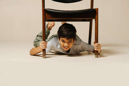 Funny indian boy playing with an armchair lying under it, holding by the legs over beige background. He is looking at the camera.の写真素材