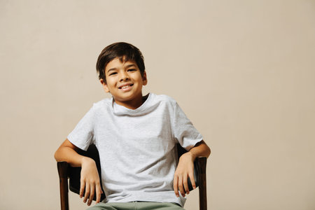Smiling indian boy sitting on an armchair in a room over beige background. He is leaning back, close...