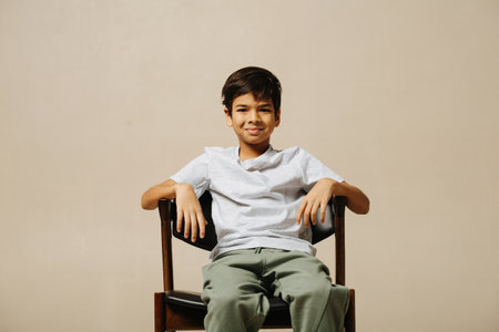 Ordinary indian boy sitting on an armchair in a room over beige background. He is leaning back, looking at the camera.の写真素材