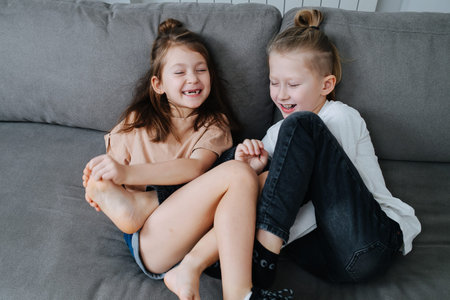 Two kids having a good time, trying to tickle each other's feet. They sit on a couch, eyes closed,...