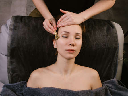 Young woman receiving temple massage in a beauty salon. She's lying on a massage bed covered with a blanket, her naked shoulders showing from under it. Hands massaging her temple with a tipped roller.の写真素材