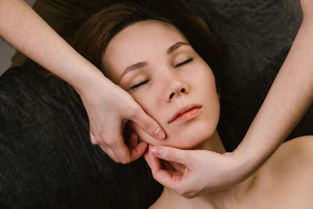 Beautiful woman receiving facial massage in a beauty salon. Her eyes closed. head and shoulders. Hand pinching her face. Diagonal framing.の写真素材
