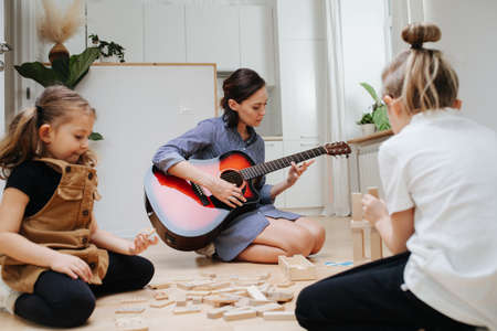 Kids busy playing on the kitchen floor with wooden blocks. While mom playing guitar in background.の写真素材