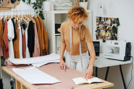 Focused seamstress checking patterns and notes, preparing to cut an unrolled fabric on the table. Meter hanging on her neck.の写真素材