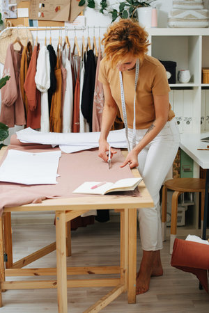 Lost in her work young seamstress cutting a fabric on the table. Meter hanging on her neck. Shelves and racks behind. She has short ginger dyed hair, looking down.の写真素材