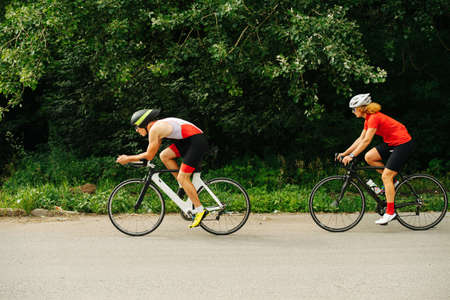 Side view of a couple riding street racing bike on a road through the park. Overgrown trees by the sides. One behind the other.の写真素材