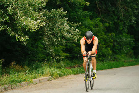 Muscular man riding his street racing bike on a road through the park. Overgrown trees by the sides. Long shot from the front.の写真素材