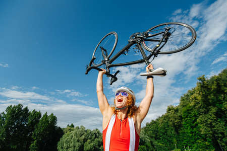 Triumphant low angle picture of a woman cyclist lifting her bike over head and yelling. Aganst sky and trees.の写真素材