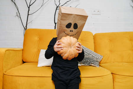 Halloween photo of a kid wearing a paper bag over his head, a scary face is drawn on it. Sitting on an orange couch, holding a pumpkin in his hands.の写真素材