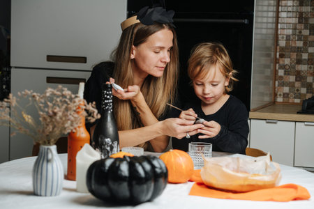 Caring mom and her little son sitting behind a round table, preparing decorations for halloween. Painting on stones, pumpkins and bottles.の写真素材