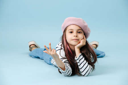 Cool fashionable tween girl in french beret lying on the floor, showing v sign. She has dyed hair strands. posing over blue background.の写真素材