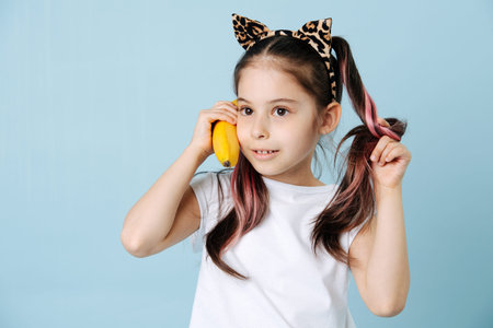 Hilarious tween girl with cat ears pressing banana to her ear as a phone. She has dyed hair strands. posing over blue background.の写真素材