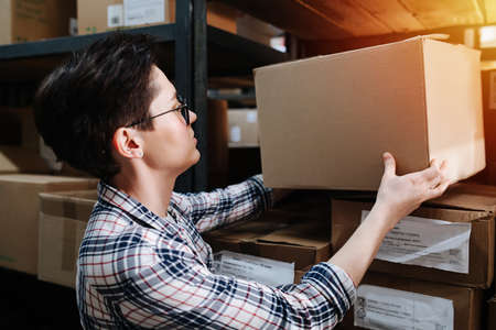 Short haired female warehouse worker placing a cardboard box on a shelf rack. She wears glasses and checkered shirt.の写真素材
