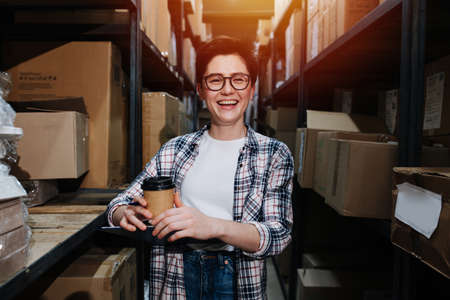 Portrait of a cheerful female warehouse worker standing between two rows of high shelves with boxes. holding a coffee.の写真素材