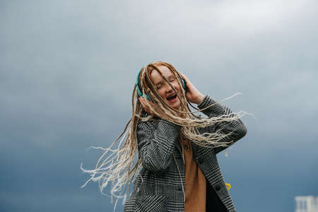 Hyped teenage girl with dreads listening music, shaking her head. upward angle. Against a gray sky.の写真素材