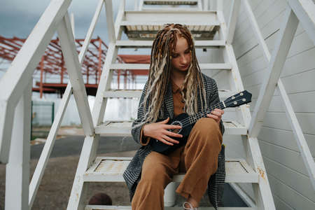 Playing a guitalele cute teenager girl with dreads sitting on a steel stairs. A gray jacket over matching brown pants and shirt.の写真素材