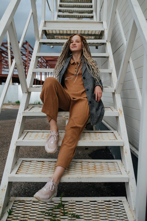 Cute portrait of a teenager girl with dreads sitting on a steel stairs. A gray jacket over matching...