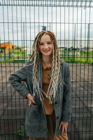 Lighthearted teenage girl with dreads standing in front of a metal fence. She is wearing a jacket....