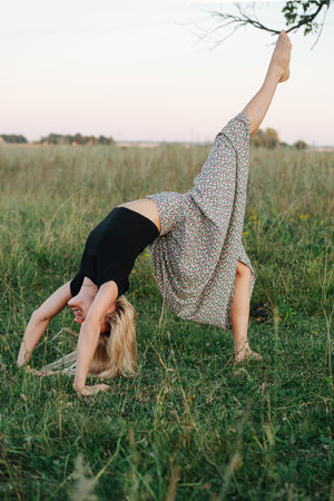 Young blond woman in a field standing in bridge pose with lifted leg. She is wearing skirt pants and black shirt.の写真素材