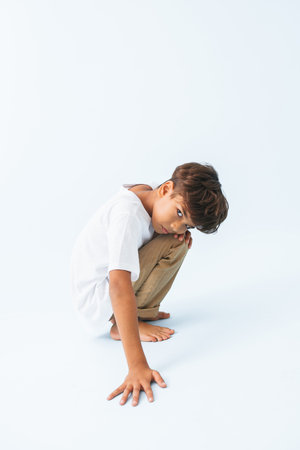 Sulking grim crouching Indian boy looking from under his brow against bluish white background. He is wearing white T-shirt. looking at the camera.の写真素材