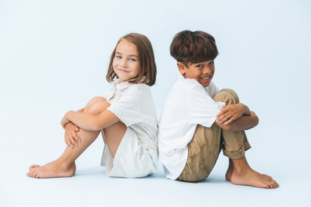 Fun portrait of awkwardly smiling boy and girl sitting back to back on the floor. against bluish white background. Both barefoot.の写真素材