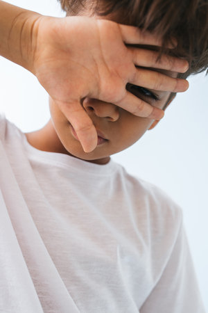 Sad Indian boy covering his face with a hand against bluish white background. He is wearing white T-shirt. looking down. close up.の写真素材