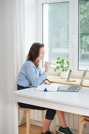 Holding a pencil business woman in blue shirt sitting behind the desk with laptop computer in home...