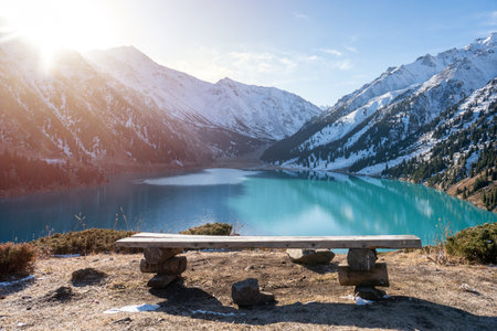Bench in front of a stunning turquoise lake surrounded by snowy mountainsの写真素材