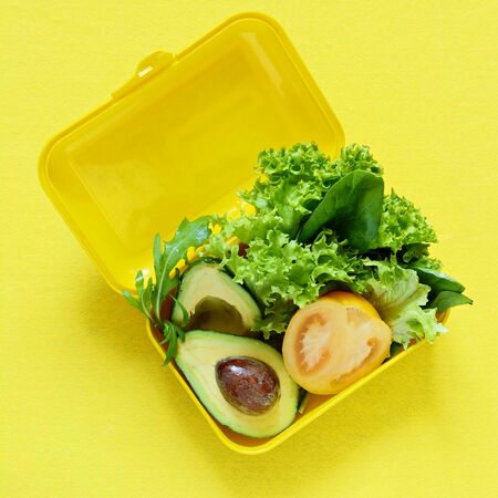 Lunchbox with a snack from lettuce, tomato, avacado on a yellow background. Healthy eating concept. Go vegan. Tasty vegetarian food in plastic box.の写真素材