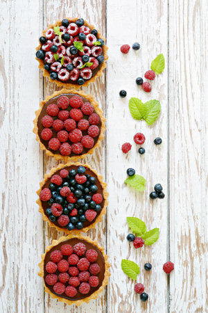 Raspberry and blueberry tartlets with chocolate ganache, fresh berries and mint leaves, selective focus. Fresh fruit tart on white background, freshly homemade fruit cake on a tableの写真素材