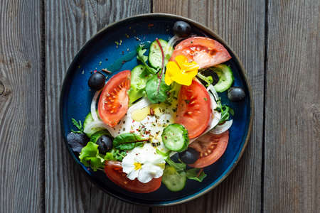 Appetizing salad with feta cheese, fresh vegetables (tomatoes, cucumbers, onions), crispy lettuce, microgreens and violet flowers. Greek salad on a wooden background. Food with flowersの写真素材