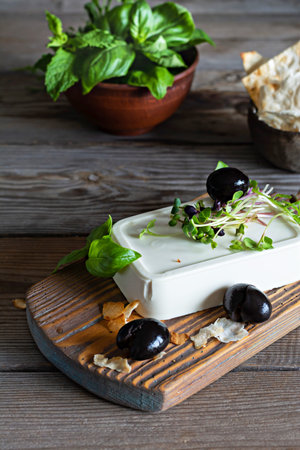 A piece of fresh feta cheese (paneer) with olives, herbs, spices and slices of flatbread (tortilla, lavash, pitta, chapatti) on a wooden board and a dark background. Greek cuisine dish, healthy food.の写真素材