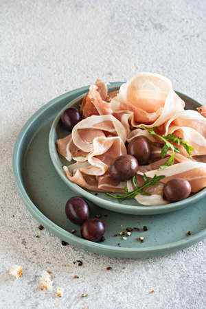 Slices of thinly sliced ham (prosciutto, Parma ham) with a bowl of olives on a light gray background. An appetizing Mediterranean snack, antipasti.の写真素材