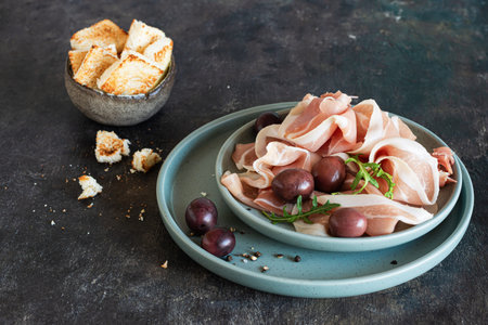 Slices of thinly sliced ham (prosciutto, Parma ham) with a bowl of olives on a dark background. An appetizing Mediterranean snack, antipasti.の写真素材