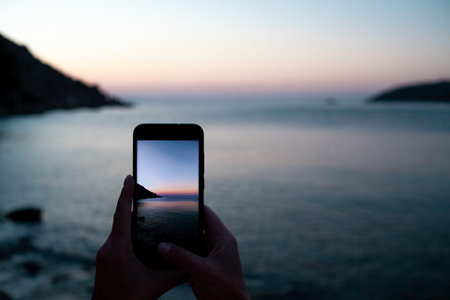 woman hands holding mobile phone at sunrise over sea bay. Taking photo of sunset over rocks in water by mobile phone. Selective focus on smartphone