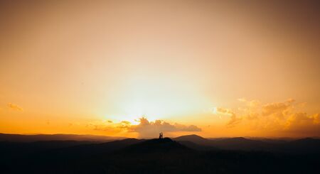 Silhouette of young couple in love enjoying a sunset over the mountains. Girl in red dress. Amazing view of young adorable couple embracing, kissing each other in the green dense mountain.