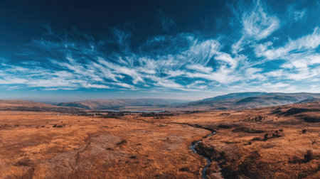 A serene panoramic view of an expansive landscape featuring a golden brown grassy field, a winding stream, and a vibrant blue sky with wispy clouds.